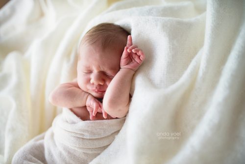 צילומי ניובורן תינוקת עטופה בד לבן בתוך שמיכה לבנה מצביעה למעלה - סטודיו אנדה יואל