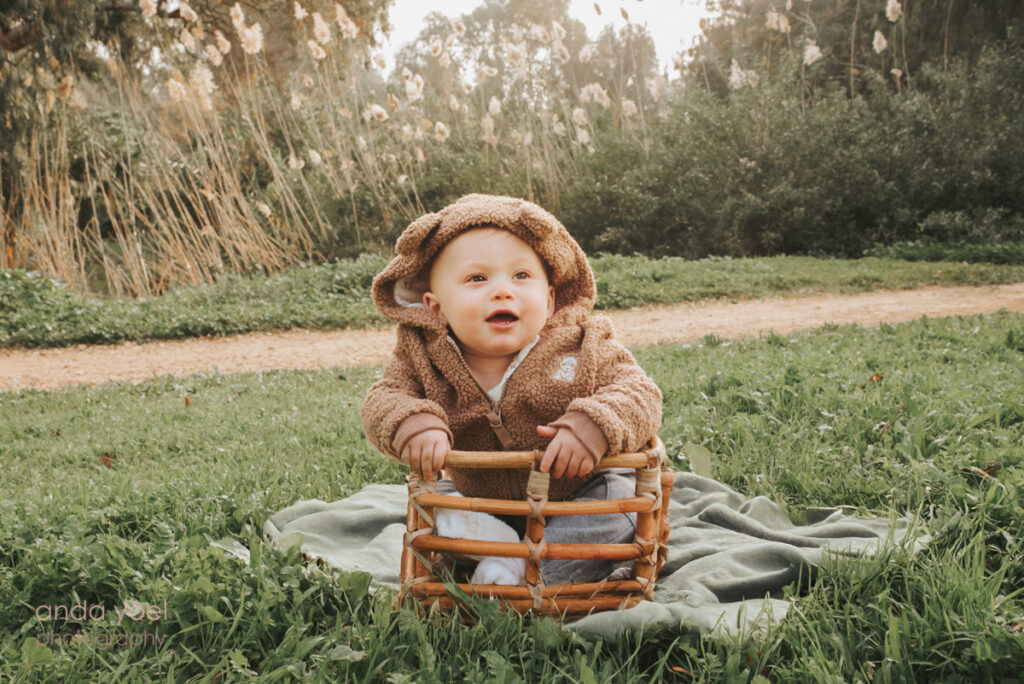 תינוק בן שנה יושב יושב בסלסלה לבוש בגדים של דובי ומחייך בצילומי גיל שנה בטבע באזור המרכז, אנדה יואל